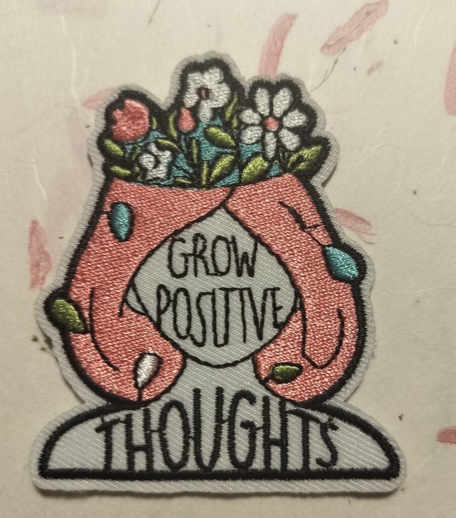 A close-up photo of an embroidered patch showing two hands cradling a head filled with blooming flowers, with the words “Grow Positive Thoughts” stitched inside. The design suggests emotional growth, gentle reprogramming, and nurturing healthier thinking patterns.
